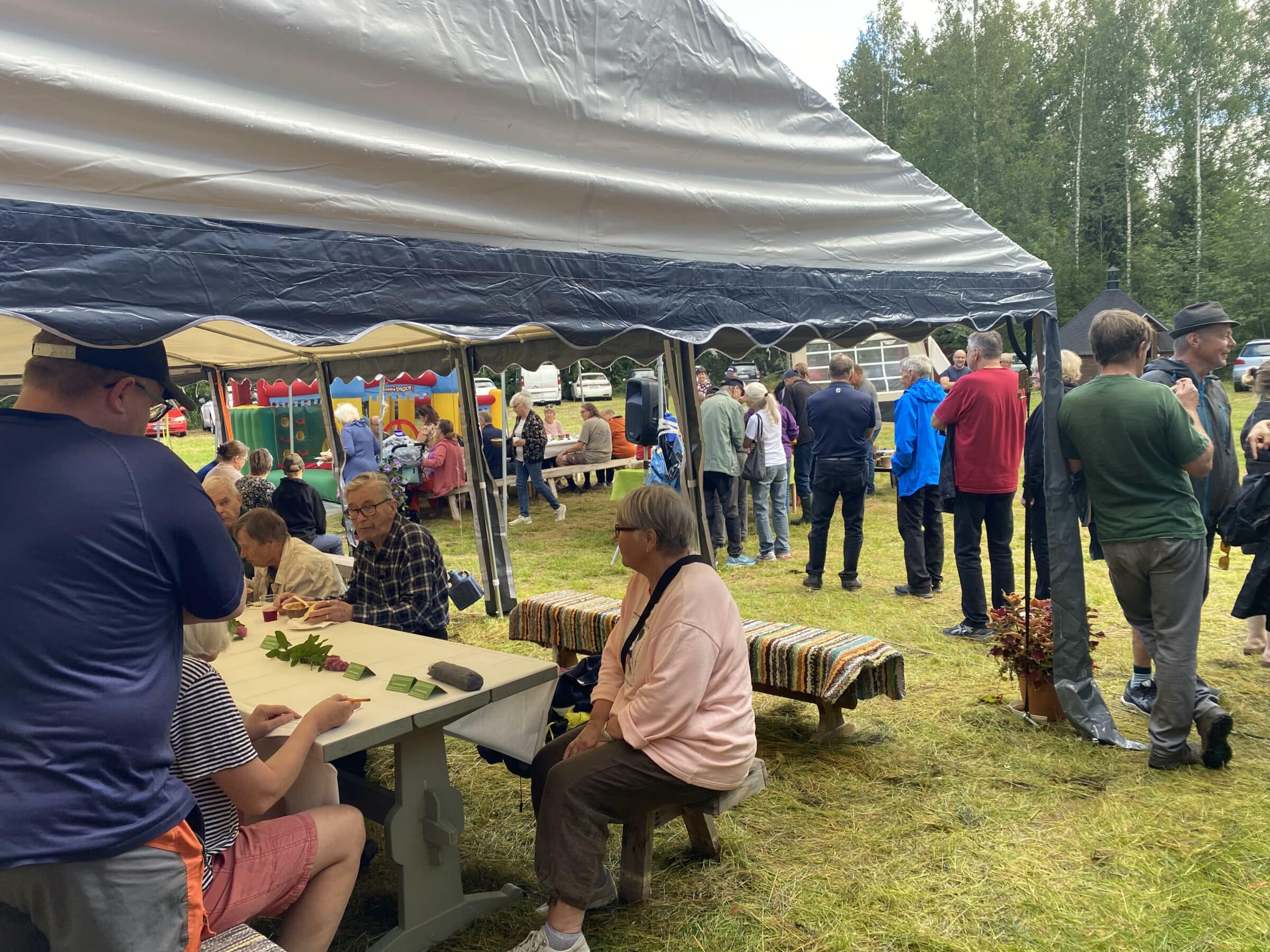 Paljon ihmisiä - osa seisoo keittojonossa ja osa istuu teltassa. Kaikki ovat ulkona. Eri värisiä takkeja.