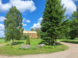 Pihalla ruohoa, puita, kiviä ja taustalla keltainen vanha maalaistalo. Tie kaartaa edustalla.