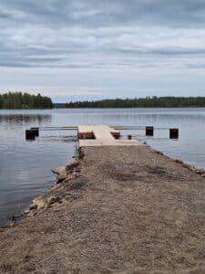 Puinen pitkä laituri jossa puomeja. Laituri on järvessä. Järvi on sininen. Laituri on puinen.