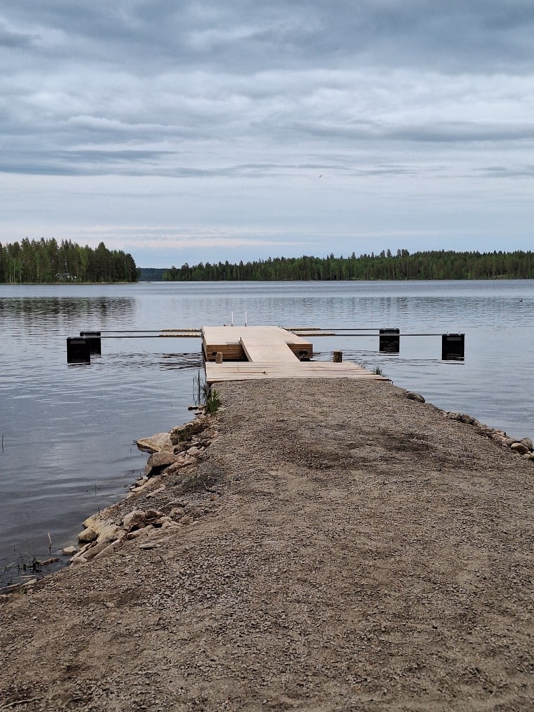 Puinen pitkä laituri jossa puomeja. Laituri on järvessä. Järvi on sininen. Laituri on puinen.