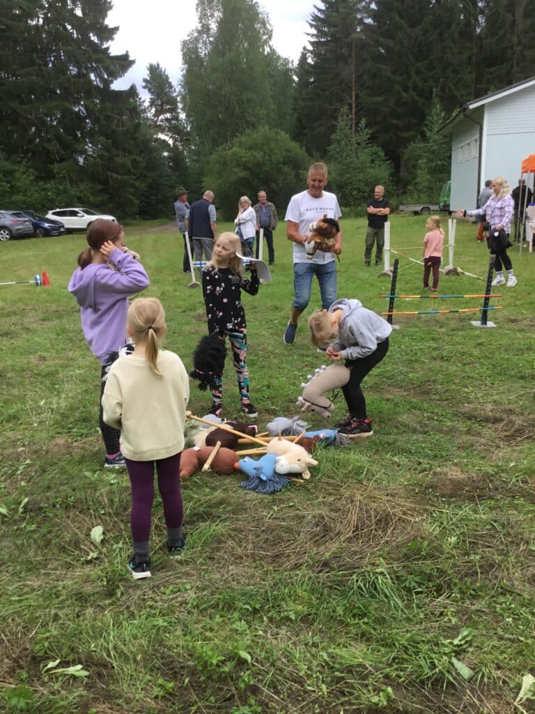 Neljä tyttöä järjestelee etualalla keppihvevosia jotka ovat maassa nurmikolla. Mies kävelee vaaleassa t -paidassa ja tuo kahta keppihevosta. Taustalla on enemmän ihmisiä.