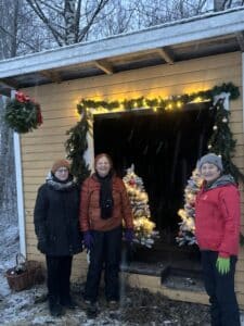 Keltaisen puisen kopin edessä Ritva Muittari, Elisa Poikolainen ja Marja-LIisa Pietiläinen. Koppi on joulukoristeltu. Valoja ja havuja. Paikka on Emos- ja Ilmolahdentien risteys.