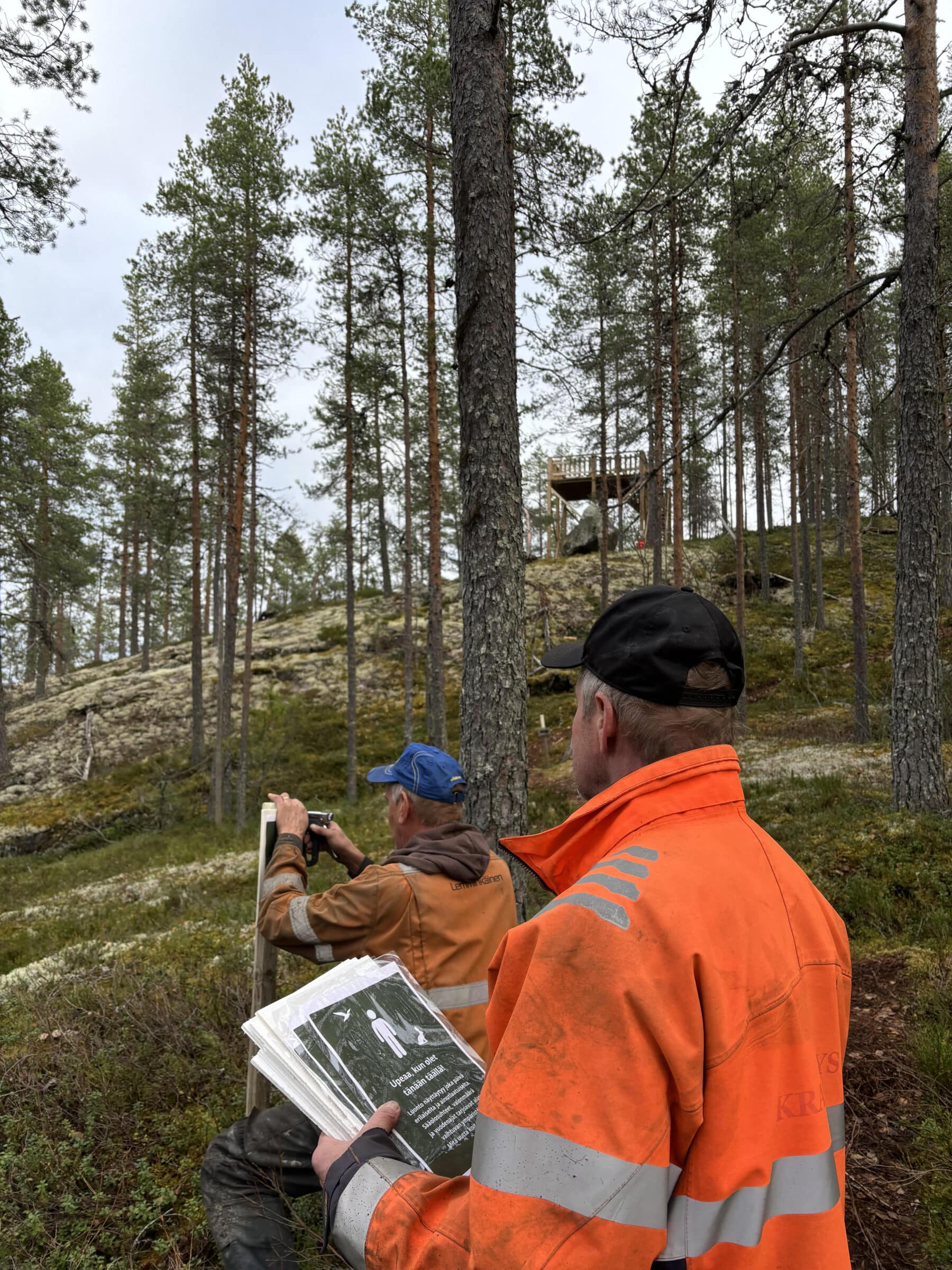 Etualalla oranssipukuinen mies jolla on vihreitä opastauluja kädessä. Toinen mies kiinnittää tauluja. Molemmat ovat metsässä. Takana näkyy järvi.